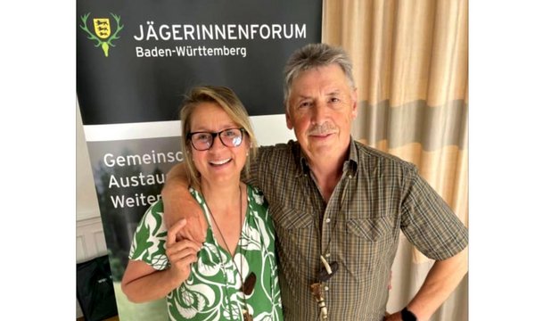 Ein Mann und eine Frau stehen lächelnd nebeneinander. Der Mann legt seinen Arm um die Frau. Im Hintergrund steht ein Schild mit der Aufschrift: "JÄGERINNENFORUM Baden-Württemberg Gemeinschaft Austausch Weiterbildung".